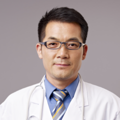 A man wearing glasses, a blue shirt, and a striped tie is dressed in a white lab coat with a stethoscope around his neck, symbolizing healthcare credentialing as he stands against a neutral background.