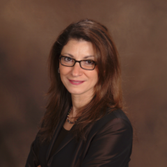 Dr. Heidi Moawad, with long brown hair and glasses, smiles warmly in a black blazer against a brown background.