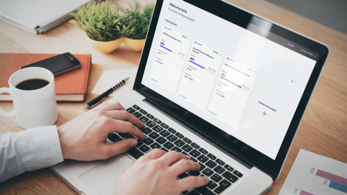 A person types on a laptop displaying a project management dashboard, analyzing medical market research surveys. A notebook, coffee cup, and plant sit on the desk.