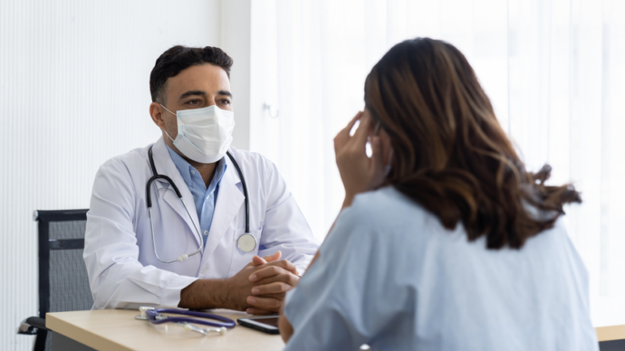 A doctor wearing a mask sits across from a patient in an office setting, discussing HIV concerns. Both individuals are engaged in conversation, and a stethoscope is on the desk.