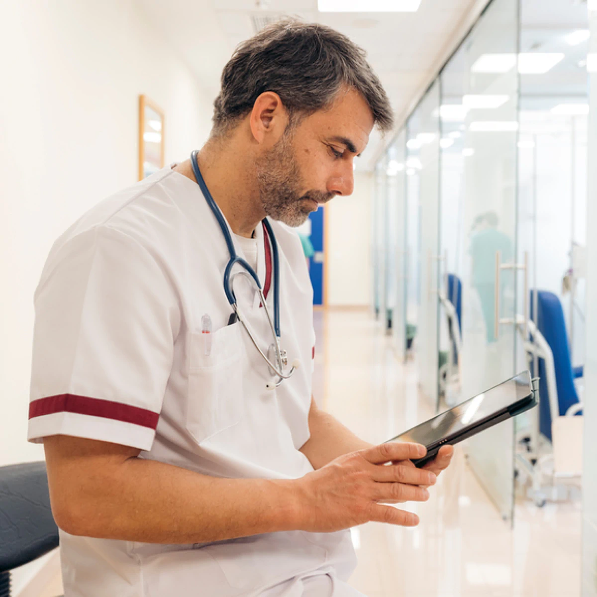 A healthcare professional in scrubs sits in a corridor, focused on his tablet.