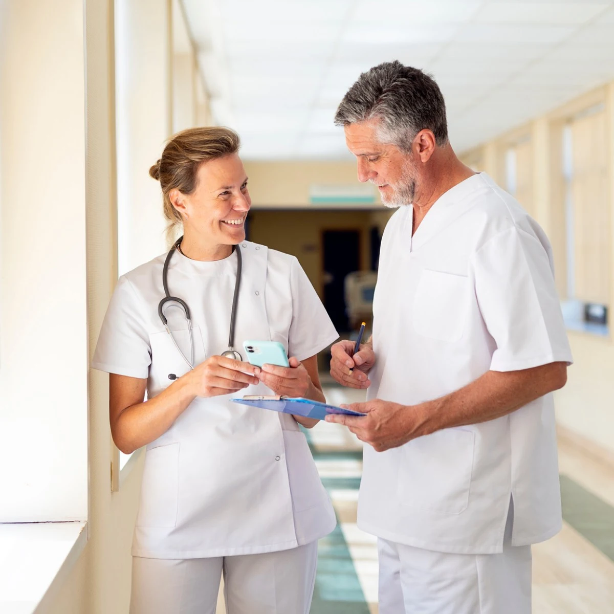 Two cardiovascular doctors in white uniforms stand in a hallway, one holding a smartphone and the other writing on a clipboard, engaged in conversation.
