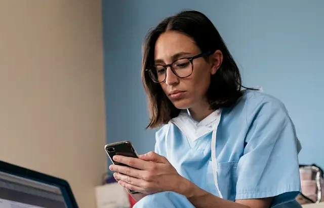 A Nurse Practitioner in medical scrubs sits by a laptop, checking their phone, with a small cup and window nearby.